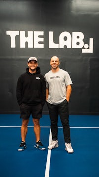 two men standing on a blue tennis court with the lab logo